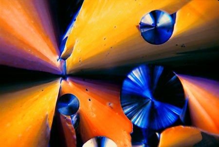 Polarized-light micrograph of broad orange crystal wedges interrupted by three dark blue spherulites, each showing a black extinction cross.