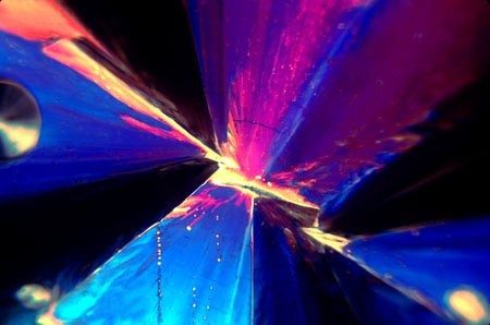 Micrograph of blue and magenta crystal blades radiating from a bright seam running diagonally across the frame.