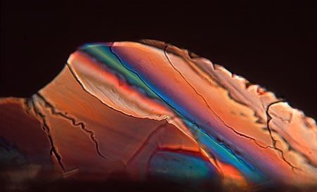 Micrograph of an orange and red banded crystal mass with a teal-green streak along one band and a cracked upper surface.