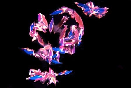 Micrograph of small pink, blue and white crystal clusters scattered in a loose spiral across a black field.
