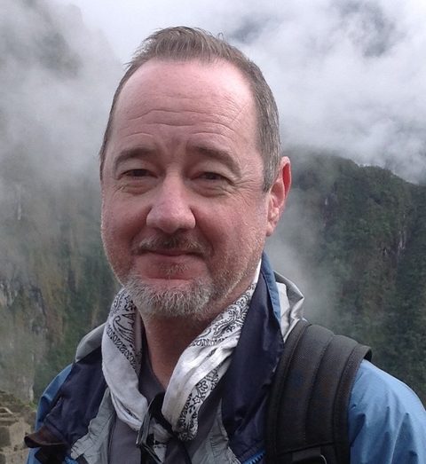 Tim Arnold outdoors in a blue jacket and scarf, misty hills behind him.
